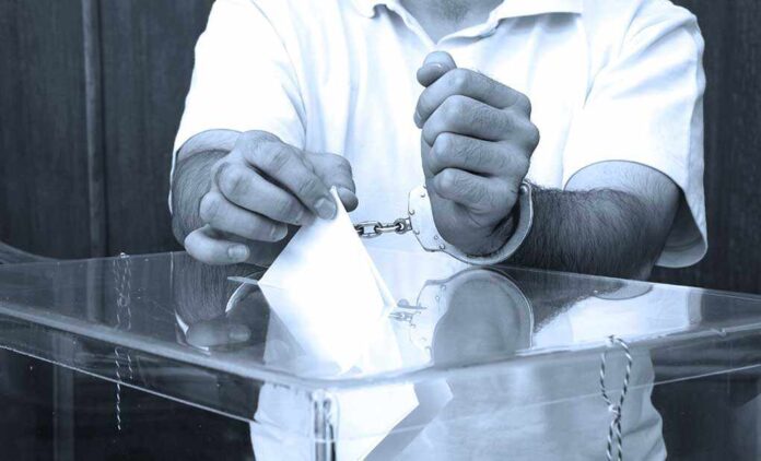 A handcuffed individual placing a ballot into a transparent voting box