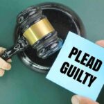 A wooden gavel next to a hand holding a blue card that says PLEAD GUILTY