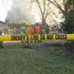 Shocking Backyard Discovery — Couple Arrested Yellow police tape marking a restricted area at a fire scene with firefighters in the background