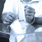 A handcuffed individual placing a ballot into a transparent voting box