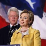 Hillary Clinton delivering a speech with Bill Clinton in the background