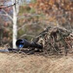 A sniper rifle partially covered in camouflage netting in a grassy area