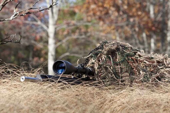 A sniper rifle partially covered in camouflage netting in a grassy area