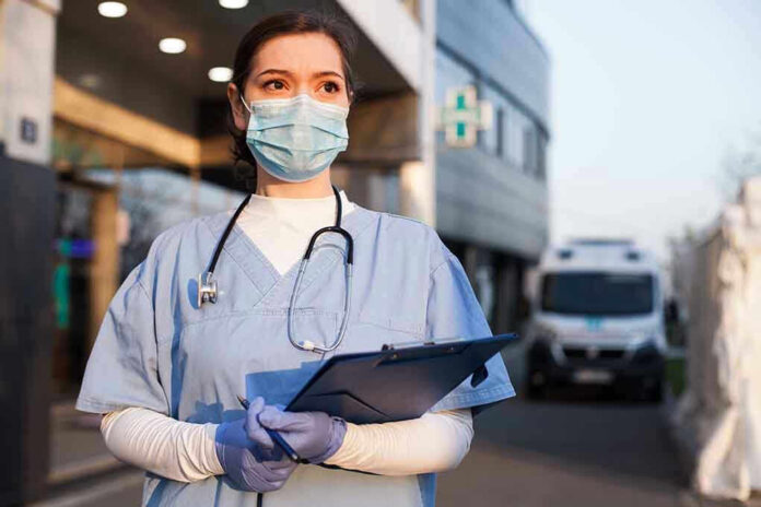 Nurse in scrubs and mask outside hospital holding clipboard.