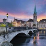 Mystery Blaze: Iconic Church in Flames Scenic view of Zurich with a bridge, historic buildings, and a sunset sky