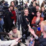 A crowded press conference with journalists and cameras surrounding a speaker