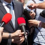 Person in suit surrounded by microphones during media interview