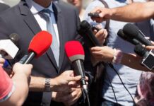 Person in suit surrounded by microphones during media interview