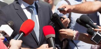 Person in suit surrounded by microphones during media interview