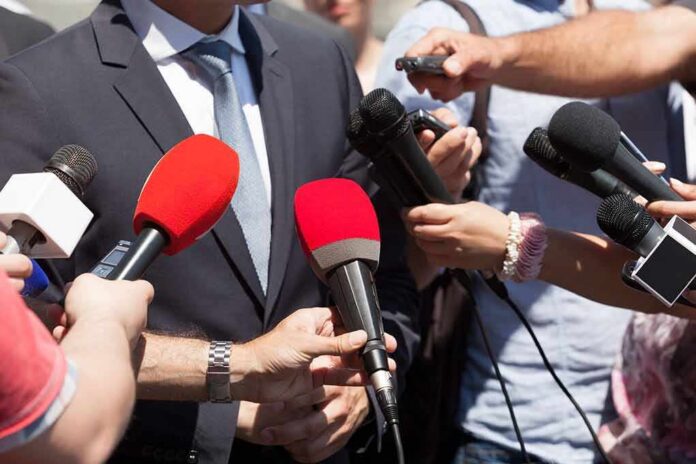 Person in suit surrounded by microphones during media interview
