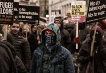 Britain Gave Citizenship to Dangerous Hamas Bodyguard Protester wearing a bandana mask amid a crowd holding signs