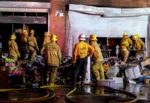 Firefighters work outside a damaged warehouse with debris and hoses