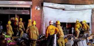Firefighters work outside a damaged warehouse with debris and hoses