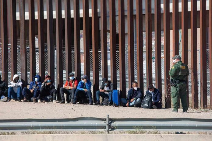 Group of people sitting by a tall border fence with an officer standing nearby